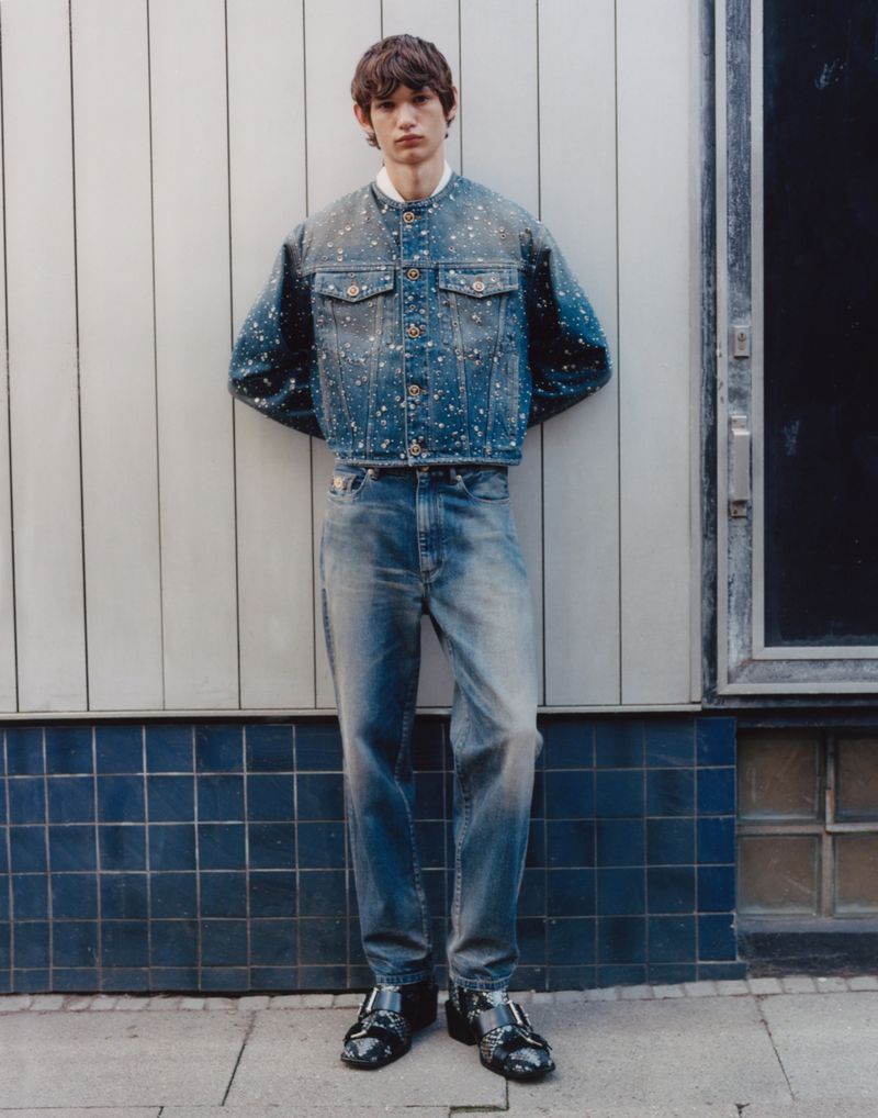 A full-length portrait of a young man in a denim jacket and jeans leaning against a white tiled wall, fashion, and urban setting.