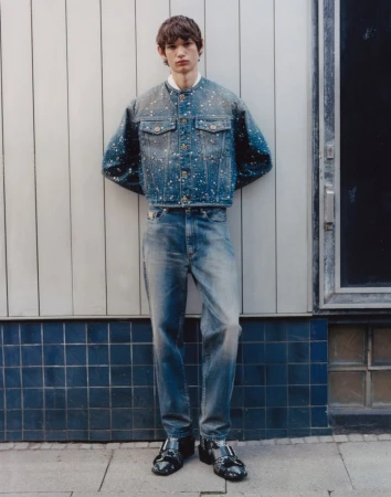 A full-length portrait of a young man in a denim jacket and jeans leaning against a white tiled wall, fashion, and urban setting.