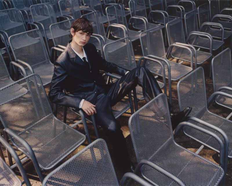 Young adult man in a black leather suit seated in a metal chair surrounded by a field of empty metal chairs outdoors.