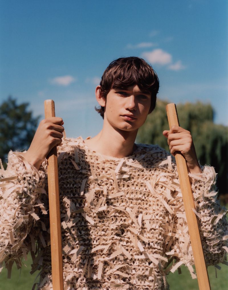 A young adult male stands outdoors in a unique textured sweater, holding wooden poles against a backdrop of blue sky and green foliage.