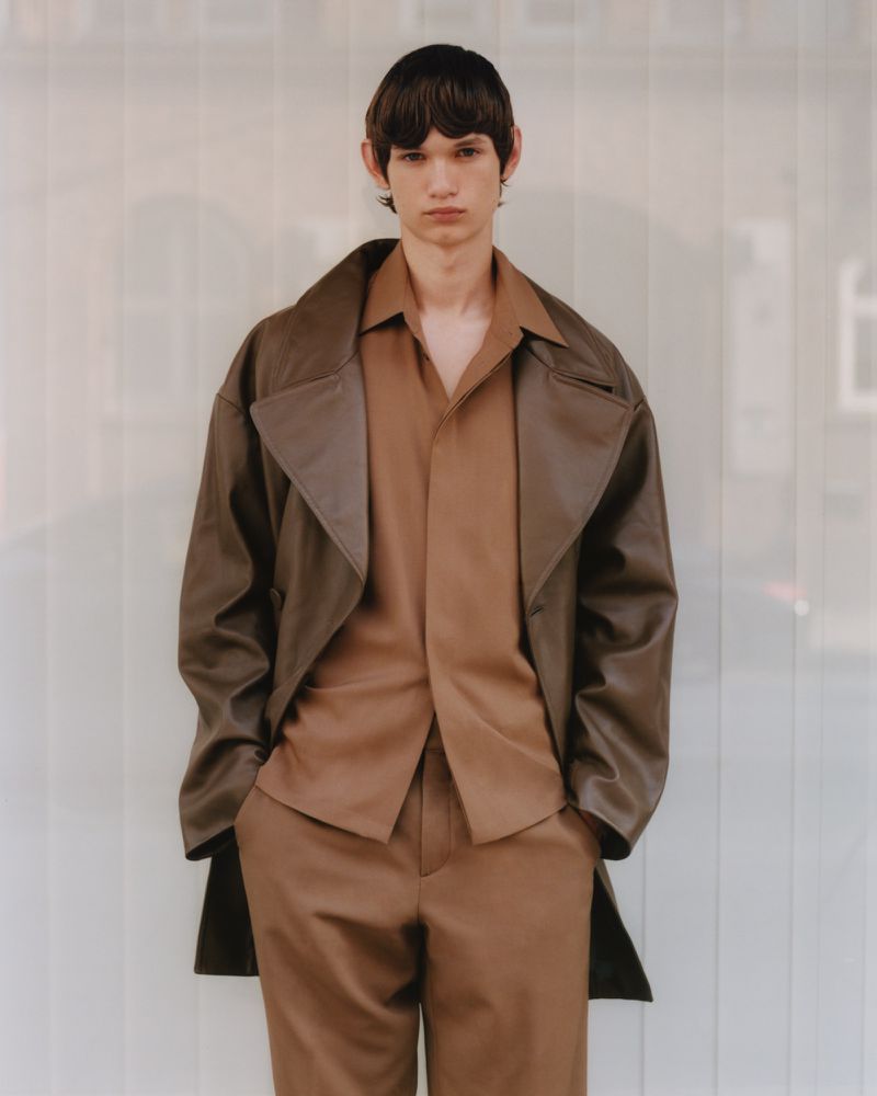 A male model poses in an indoor setting, wearing a brown leather trench coat, a brown button-down shirt, and brown pants, looking directly at the camera.