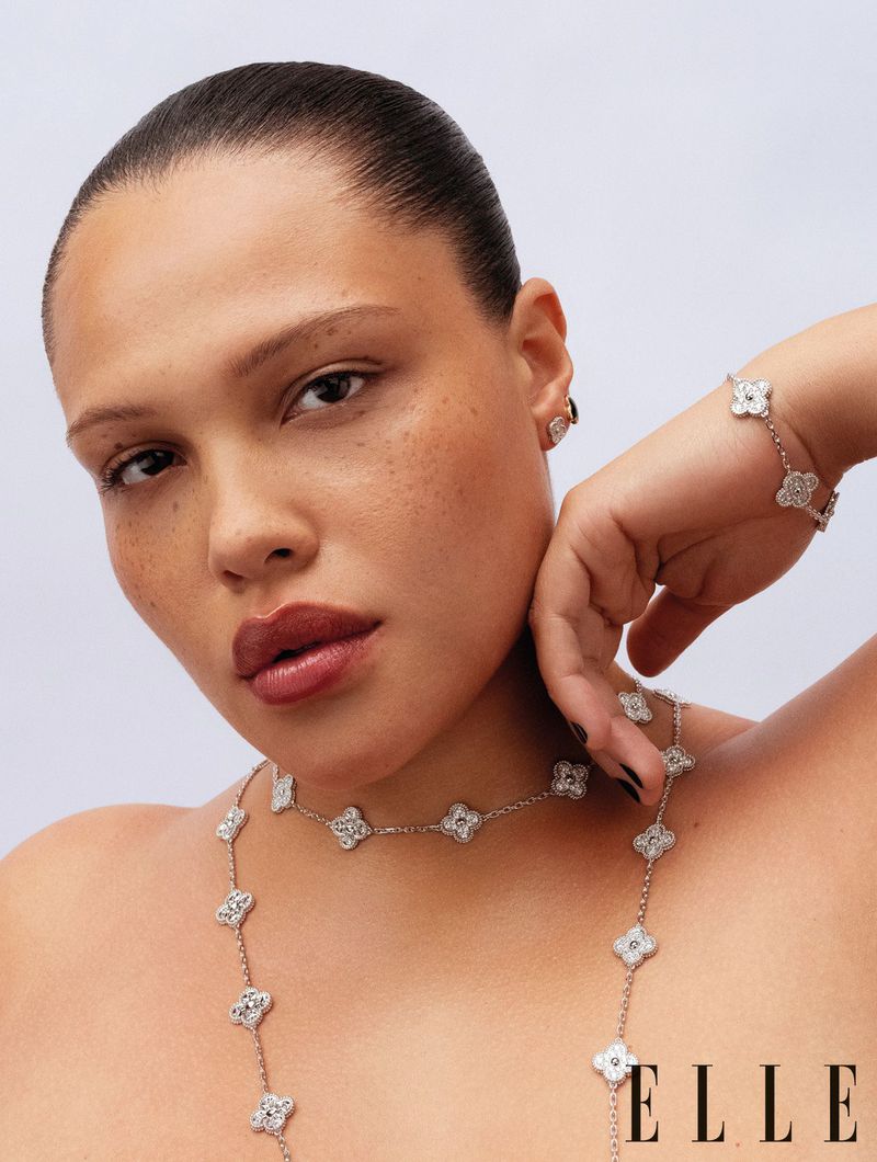 Close-up studio shot of a woman with a slicked-back bun, wearing a necklace, bracelet, and earrings with flower-shaped pendants.