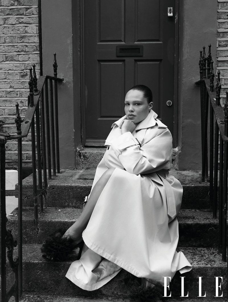 Black and white fashion portrait of adult female sitting on outdoor steps, wearing trench coat with dark door and brick wall backdrop.