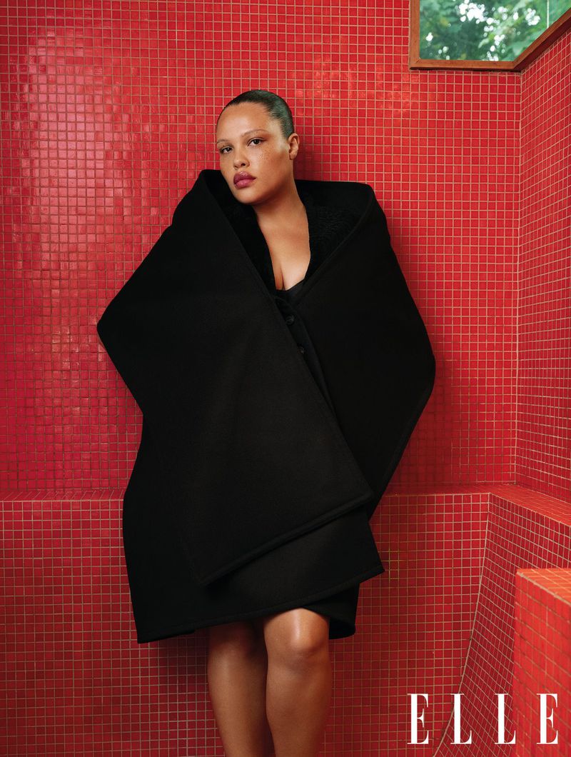 Adult female poses in a black coat against a red tiled wall indoors, fashion editorial shot, window light, modern style.