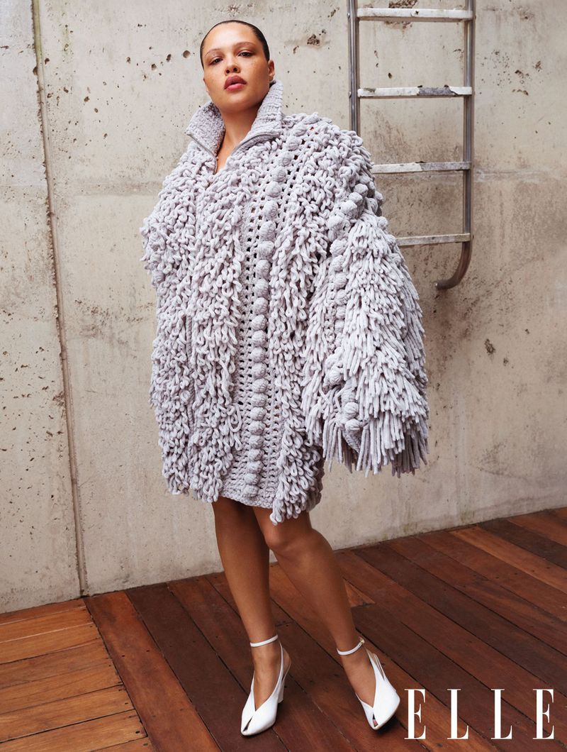 An adult female poses in a studio wearing a grey, fringe-textured jacket dress and white high heels against a concrete background.