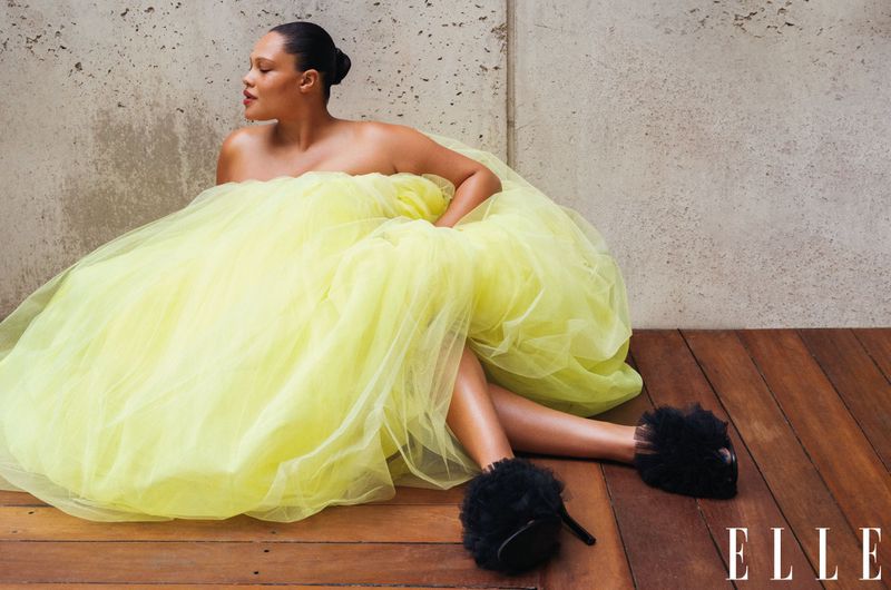 Woman in a yellow tulle dress sits on the wooden floor against a concrete wall. She wears black heels with furry accents and poses.