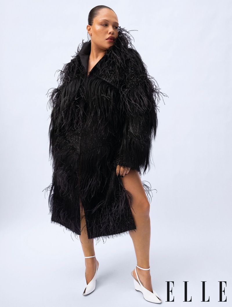 Woman in black feathered coat poses in a fashion shot against a white backdrop, wearing white heels with the ELLE logo in the corner.