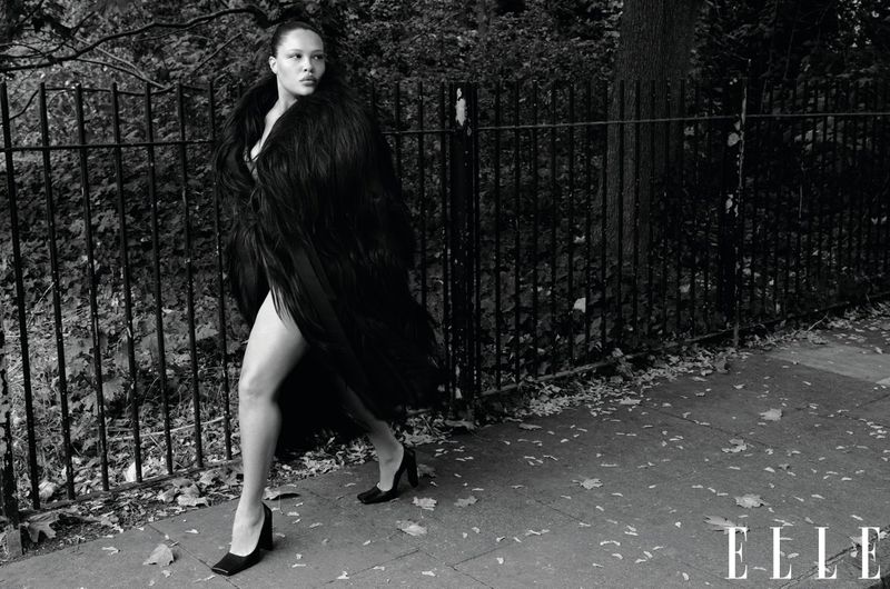 Adult woman walks on a sidewalk beside a metal fence in a black furry coat and black heels in this outdoor, black and white fashion shot.
