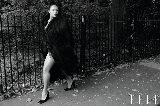 Adult woman walks on a sidewalk beside a metal fence in a black furry coat and black heels in this outdoor, black and white fashion shot.