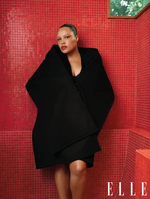 Model Alva Claire wearing a heavy black coat standing against a red tiled wall for Elle UK