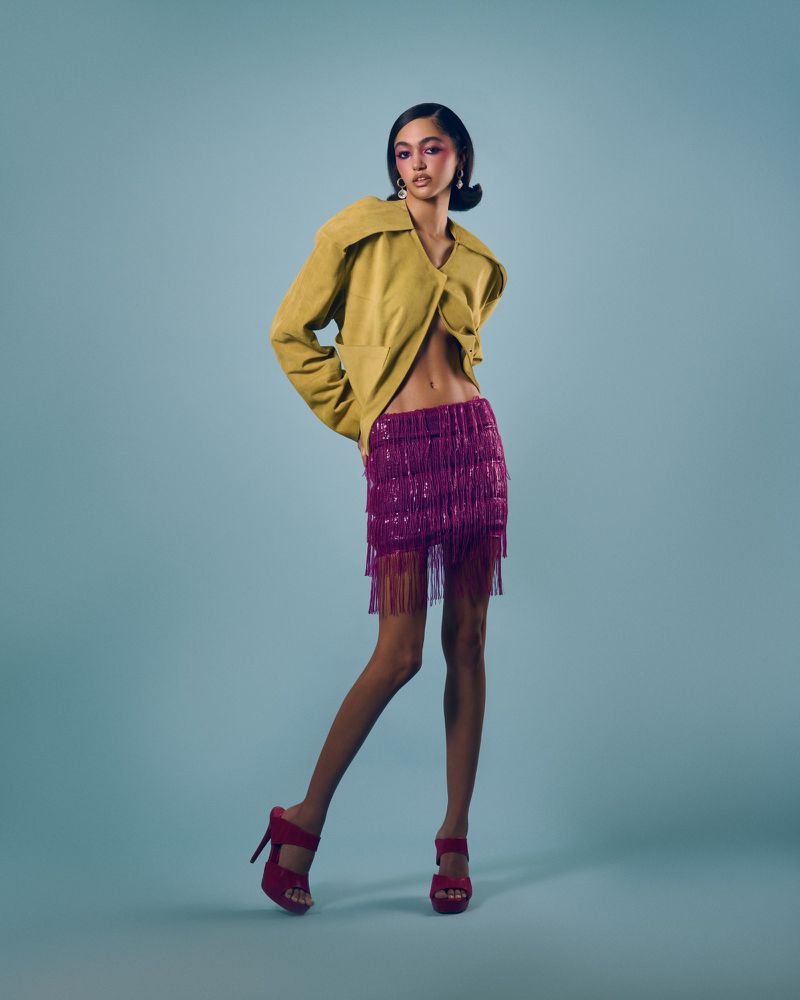 Full-length portrait of a brunette woman wearing a yellow jacket, purple sequin skirt, and pink heels in a studio setting.