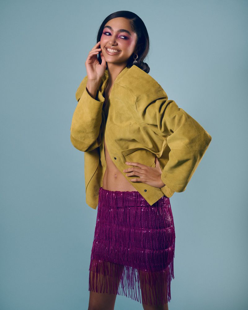 Smiling young woman with a yellow jacket and magenta fringe skirt poses in a studio with a light blue background, looking at the camera.