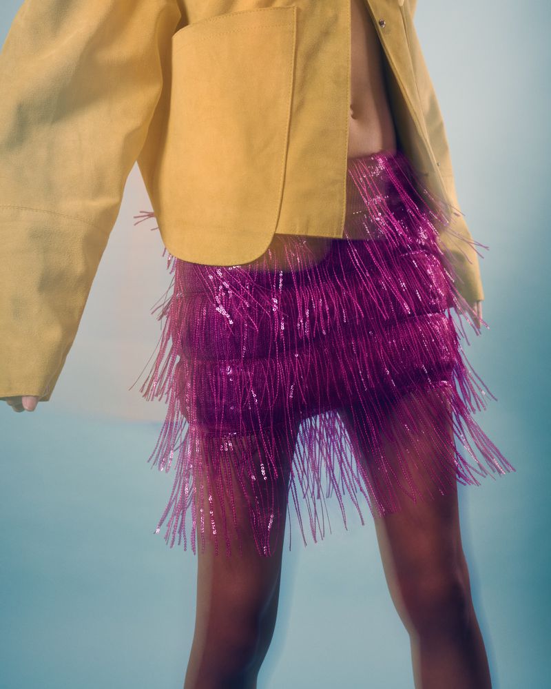 Woman poses wearing a yellow jacket and a layered, pink, fringe skirt against a blue background in a fashion forward studio shot.