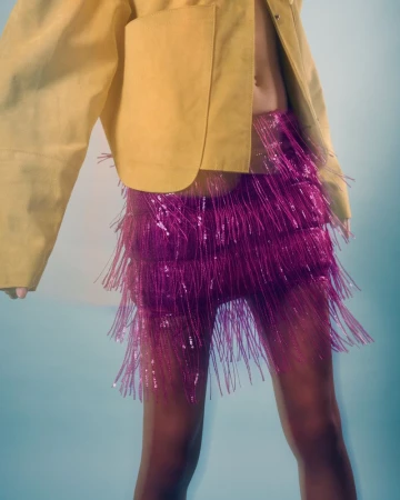 Close-up of a person wearing a yellow jacket and a vibrant magenta fringed skirt against a blue background.