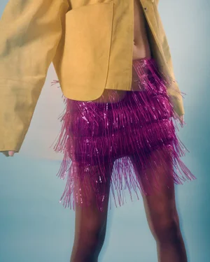 Close-up of a person wearing a yellow jacket and a vibrant magenta fringed skirt against a blue background.