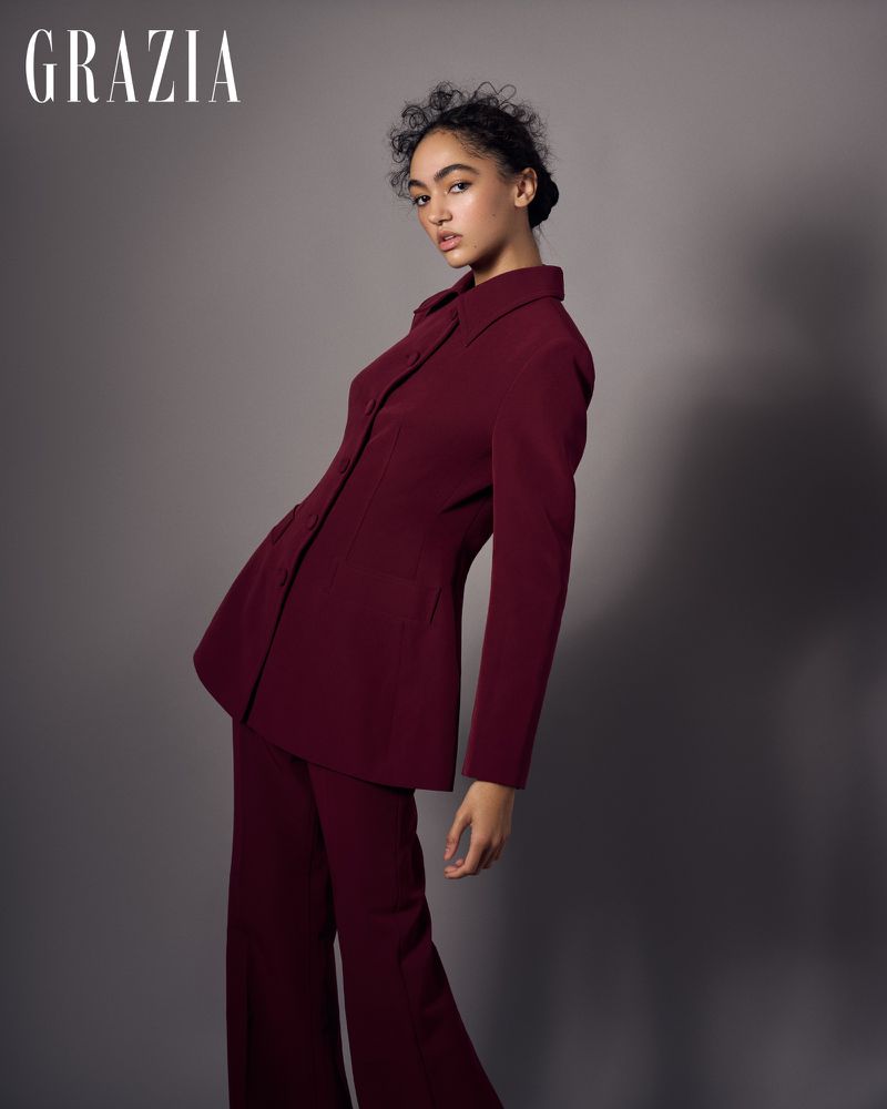 Fashion editorial shot: Woman in a maroon suit poses in a studio with a gray background, showcasing modern and elegant style in portrait.