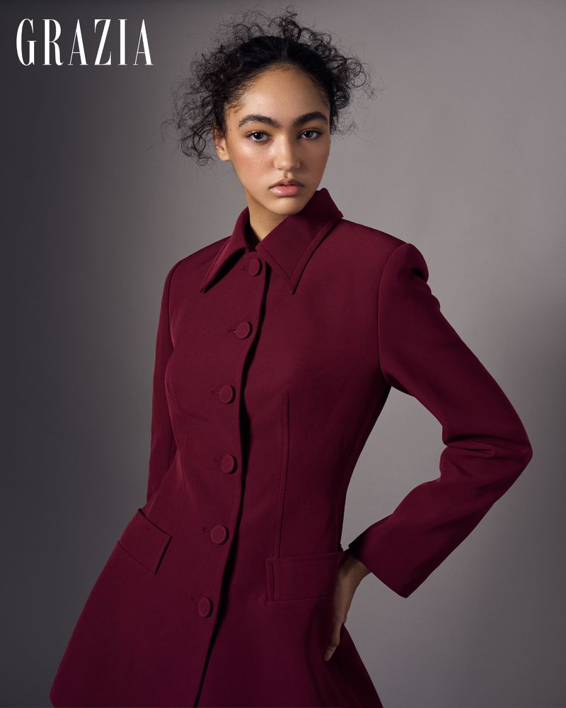 Elegant portrait of a young woman in a fitted burgundy blazer dress with dark curly hair, posing indoors against a neutral grey backdrop.