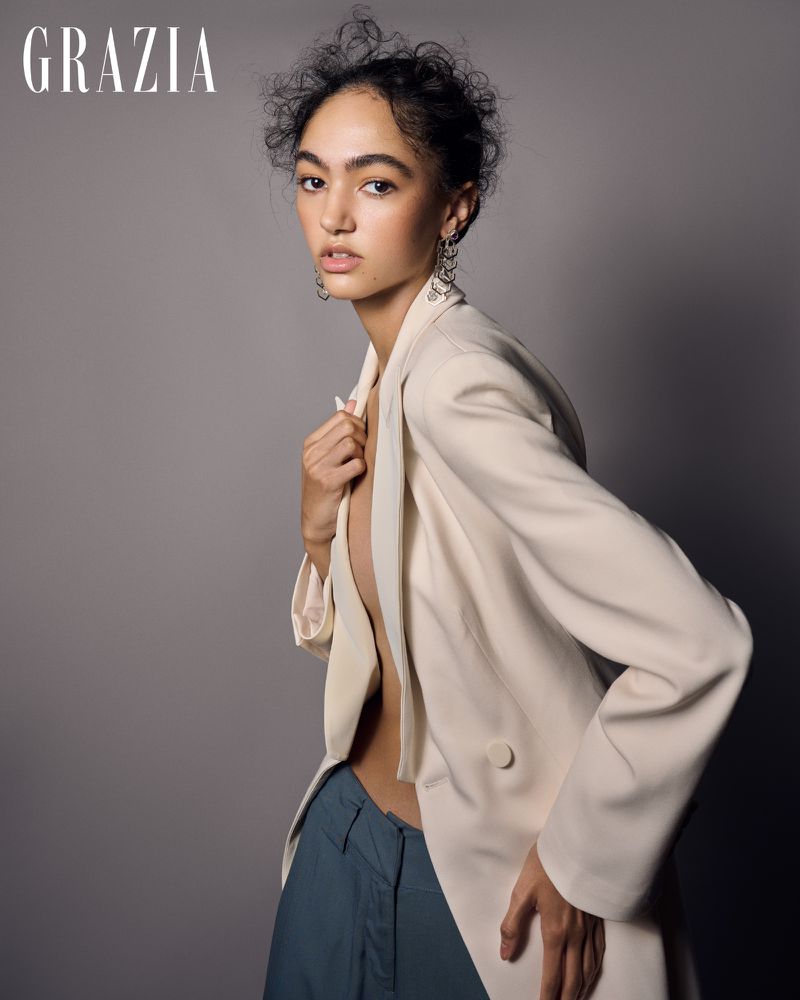 Fashion portrait of a woman with curly hair wearing a blazer and trousers in a studio with gray background, posing and looking at camera.