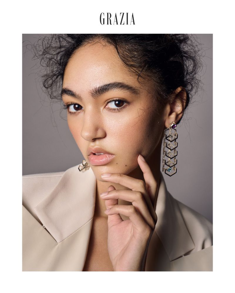 Close-up of an adult female fashion model in a studio wearing a neutral-toned blazer and geometric earrings looking at the camera.