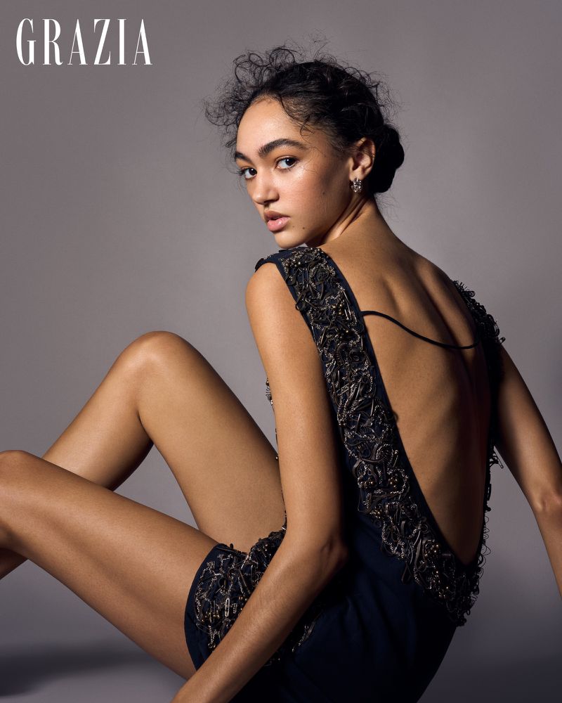 Portrait of a woman with dark hair wearing a low back dress sitting in a studio setting. The model gazes at the camera.