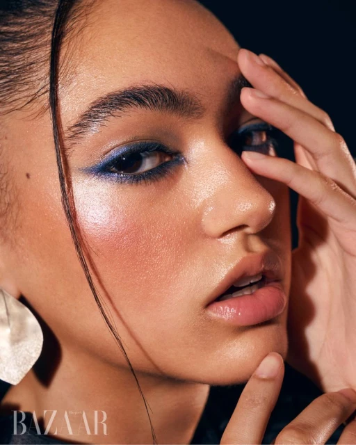 Close-up beauty shot of a young adult woman with glossy skin, blue eyeliner, and silver earring posing in front of a dark background.