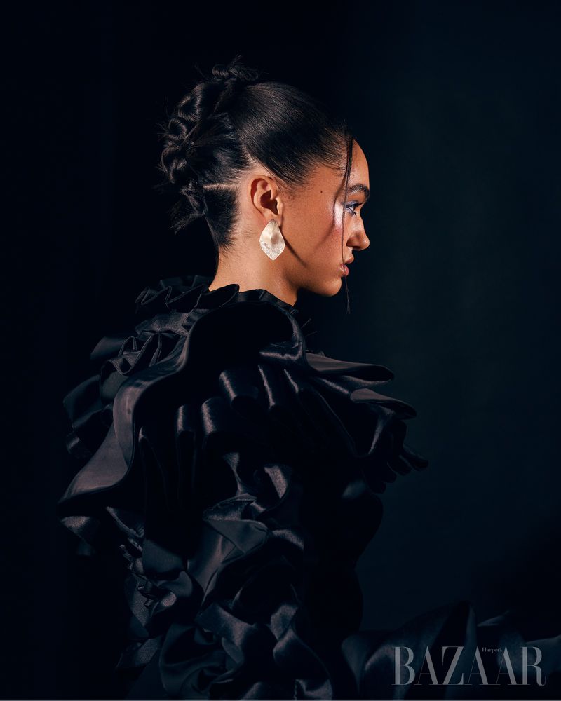 Glamour portrait of an adult female in profile wearing a black ruffled dress and statement earrings against a black backdrop.