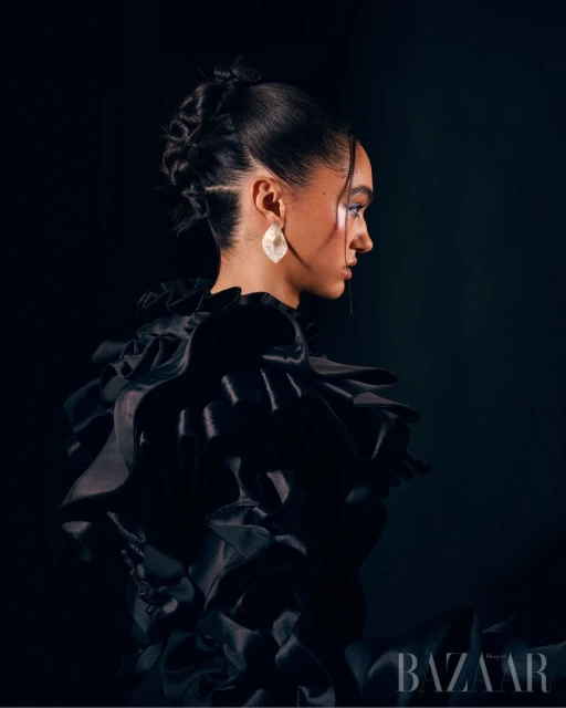 Glamour portrait of an adult female in profile wearing a black ruffled dress and statement earrings against a black backdrop.