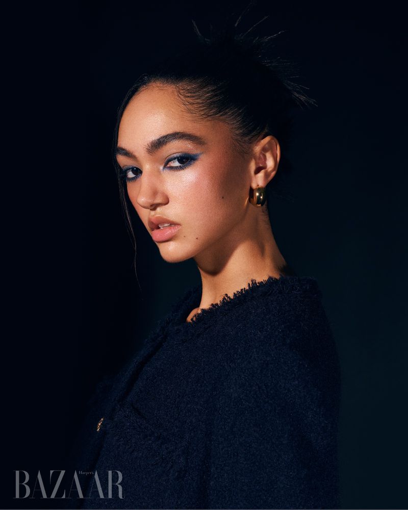 Close-up portrait of a woman with dark hair pulled back and blue eye makeup, wearing a textured navy jacket in a dark studio setting.