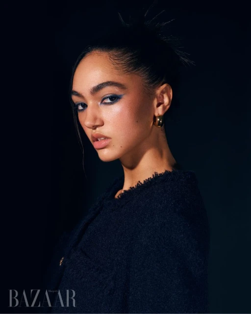 Close-up portrait of a woman with dark hair pulled back and blue eye makeup, wearing a textured navy jacket in a dark studio setting.