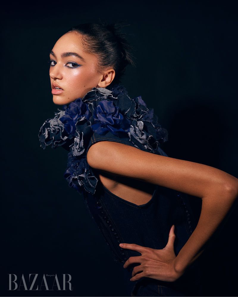 Fashion portrait of a woman with dark hair and makeup, wearing a dark blue dress with flower embellishments against a dark background.