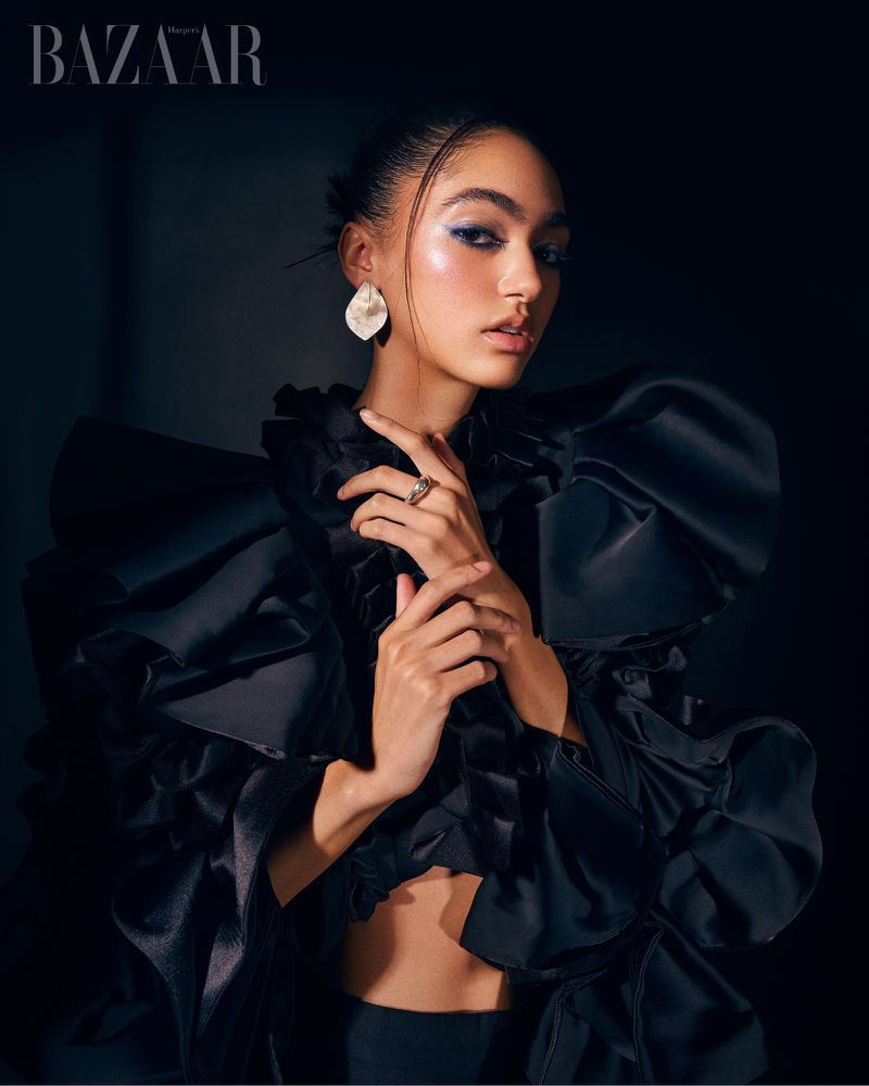 Fashion portrait of a model in a black ruffled top with blue eye makeup and white earrings against a dark backdrop in studio light.