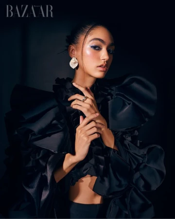 Fashion portrait of a model in a black ruffled top with blue eye makeup and white earrings against a dark backdrop in studio light.