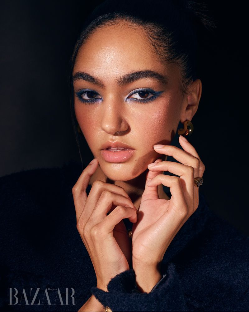 Close-up of a woman with makeup and dark hair, gazing directly at the camera in a studio setting, wearing a dark top.