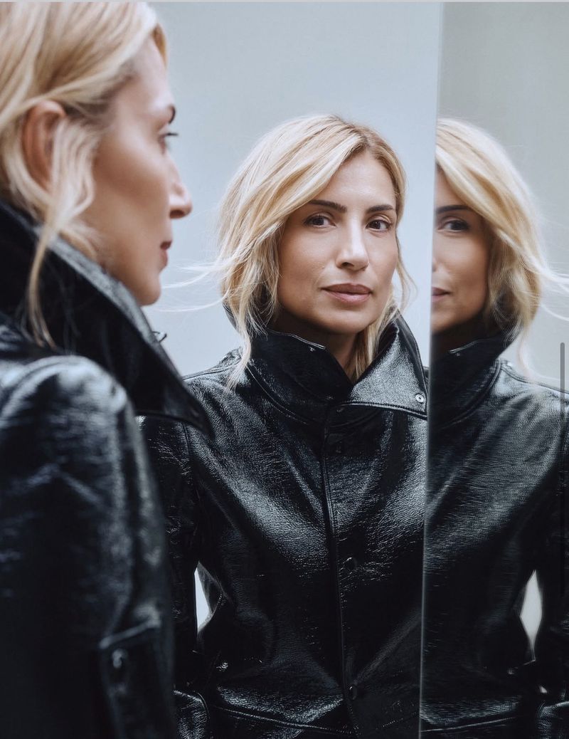 Blonde woman in shiny black jacket poses indoors with reflection in mirror. Portrait highlights fashion and modern studio setting.