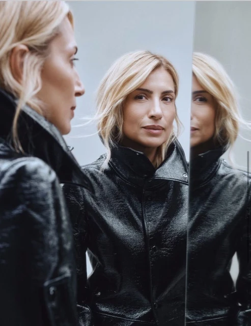 Blonde woman in shiny black jacket poses indoors with reflection in mirror. Portrait highlights fashion and modern studio setting.