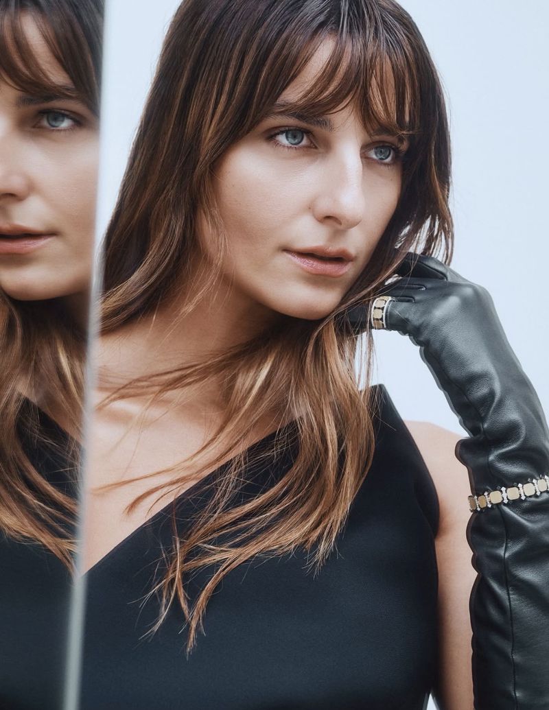 Elegant woman with bangs in black dress and leather gloves poses in front of a mirror, creating a reflected image in a light studio setting.
