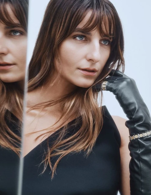 Elegant woman with bangs in black dress and leather gloves poses in front of a mirror, creating a reflected image in a light studio setting.