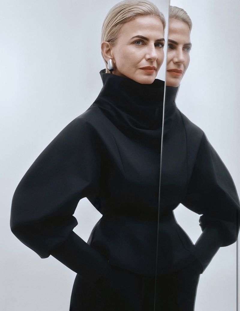 Elegant adult woman in a black dress, posing with a mirror reflection against a plain light background in a portrait shot.