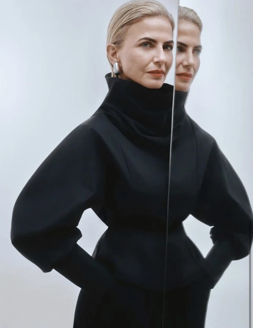 Elegant adult woman in a black dress, posing with a mirror reflection against a plain light background in a portrait shot.