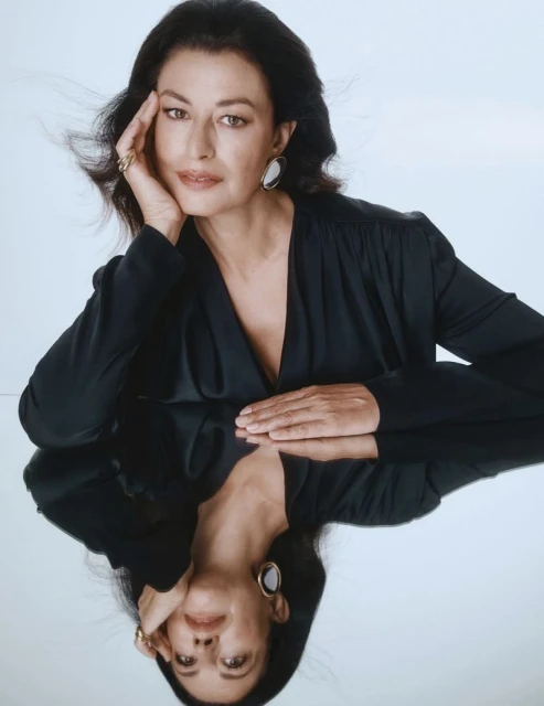 Elegant portrait of a mature woman in a black dress with jewelry, resting her head on her hand, reflected on a glossy surface.