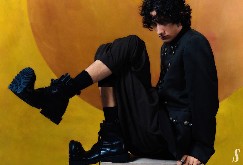 Male model with curly hair poses in black jacket, pants, and boots against a yellow background in a fashion studio shot.