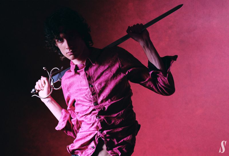 Young adult with curly hair holds a sword on his shoulder, wearing a textured button-down shirt, posed against a diffused pink backdrop.