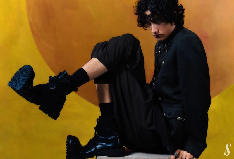 A young man in a dark military-style jacket and chunky black boots seated against a warm yellow backdrop