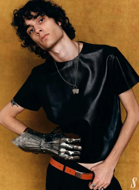 Musician Sombr wearing a black leather shirt and a metal knight gauntlet against a warm textured gold studio backdrop.
