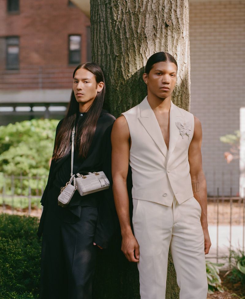 Two fashion models pose in front of tree and brick building. One in white vest and pants, one in black coat. Neutral tone fashion shoot.
