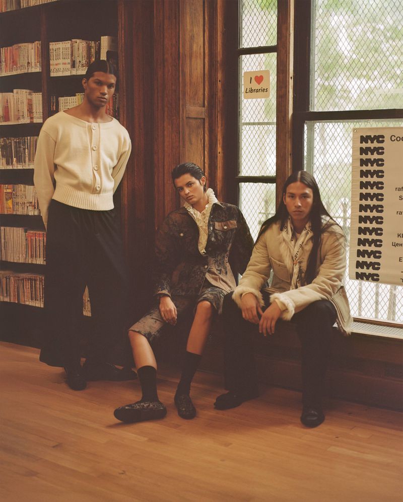 Three people in a library setting; a tall man in a white sweater stands while the other two sit on a wooden bench by a window.