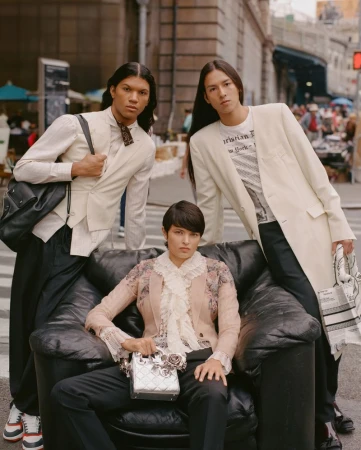 Three fashion models showcase designer clothing and accessories while posing on a black couch in an urban outdoor setting.