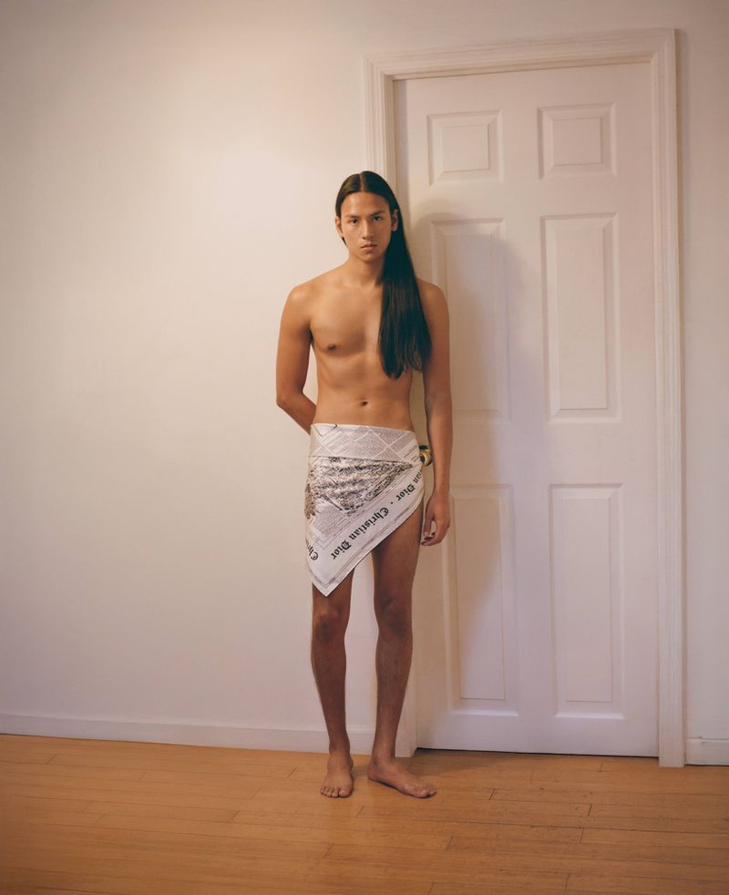 Full shot of a topless adult male with long hair wearing newspaper as a skirt, posing in front of a white door and wall.
