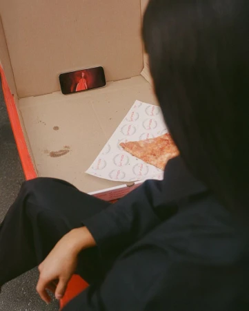 Overhead view of a person seated watching an on-screen figure in red, with pizza and a pizza box visible indoors.