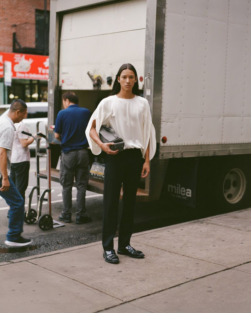 A model in a white blouse and black pants stands on a city sidewalk next to a moving truck with workers in the background.